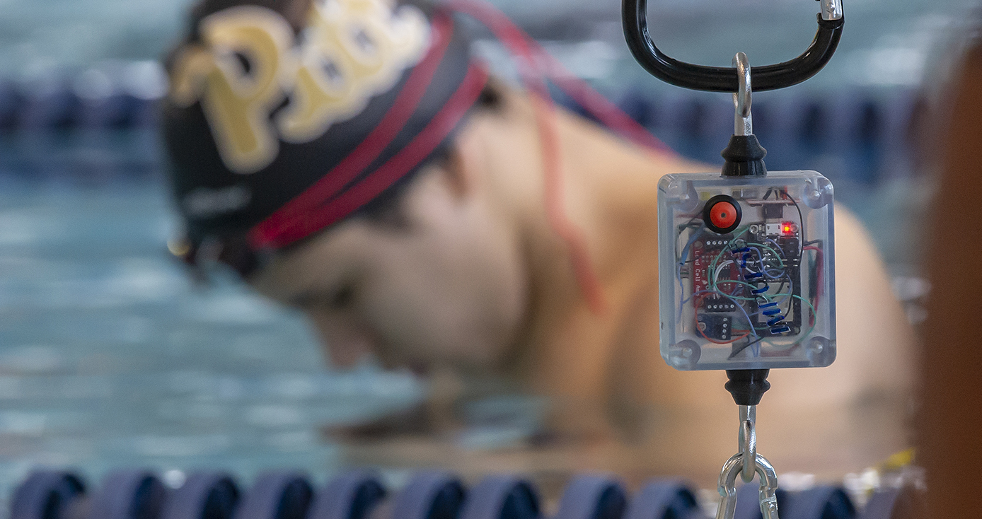 An electronic device hanging by a clip with a swimmer with a Pitt cap on in the background