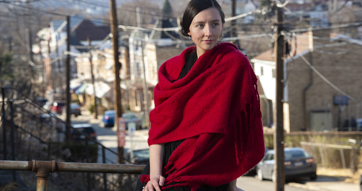 Howell draped in a red scarf, sitting on a porch ledge with a city street in the background