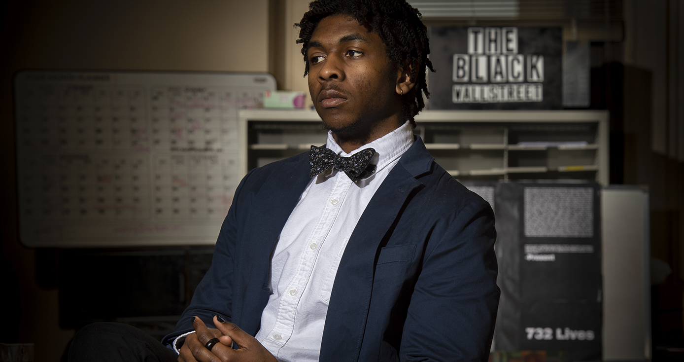 Kent-Smith in a jacket, white shirt and bowtie, sitting pensively in front of a sign that says "the black wallstreet"