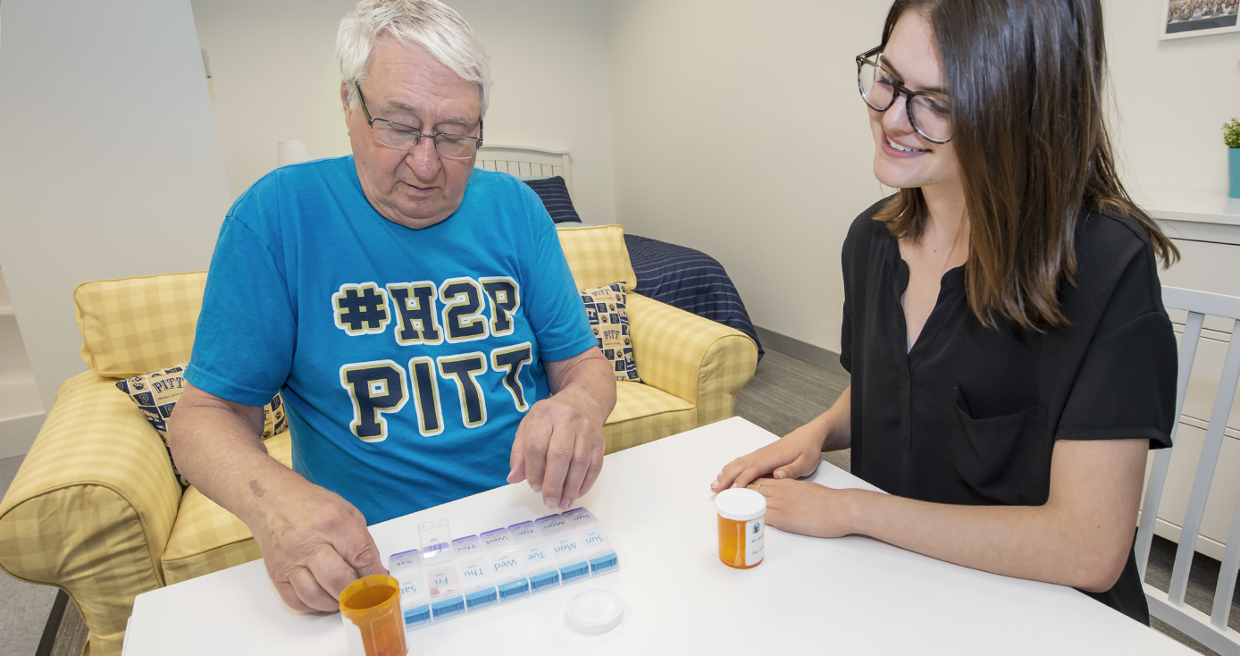 Dennis Stark, in light blue Pitt T-shirt sitting in a plaid yellow armchair, placing pills into a pill counter placed on a white table.  Samantha Whitney sits at the table with him, in black polo top, observing. Prescription pill bottles are on the table.