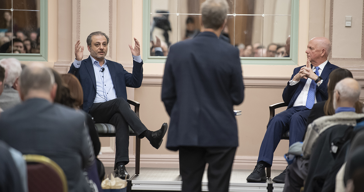 Preet Bharara and David Hickton seated, facing toward an audience; back of audience's heads are visible. Bharara gestures while speaking. Hickton looking toward Bharara.