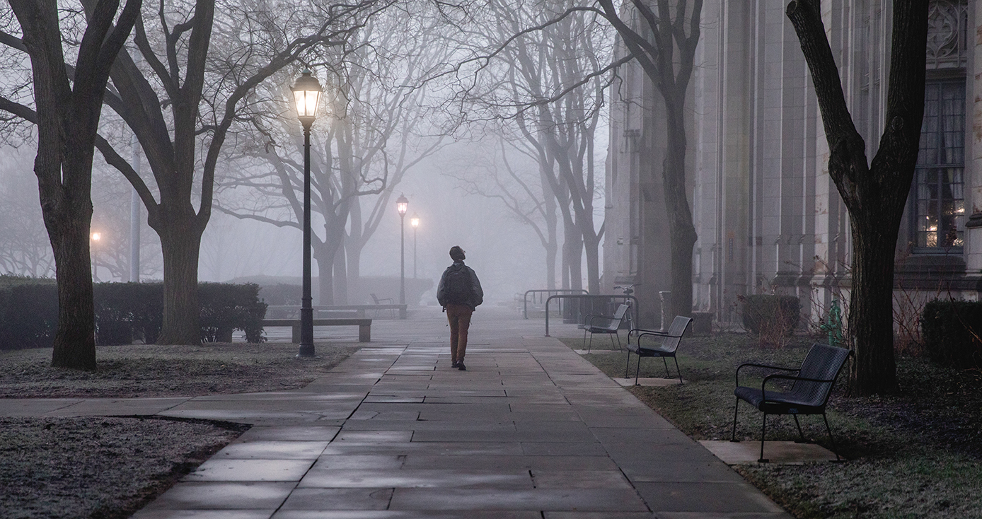 A person walks alone on a sidewalk in foggy weather beneath street lamps