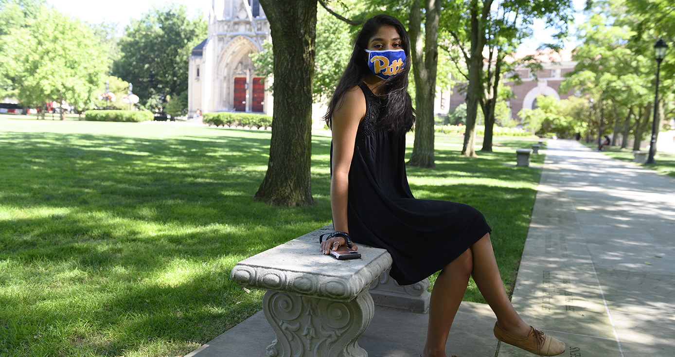 a woman in a black dress wearing a Pitt branded mask and sitting on a bench outside Heinz Chapel