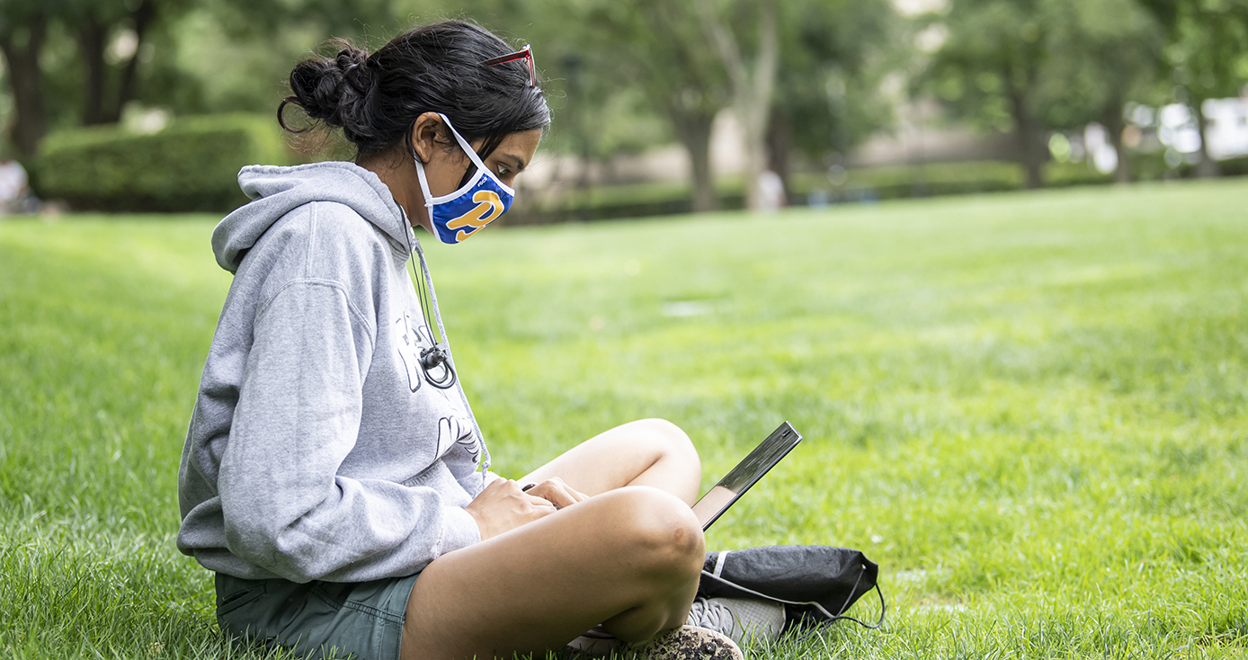 A student in a gray sweatshirt and a blue Pitt face mask works on a laptop in a field