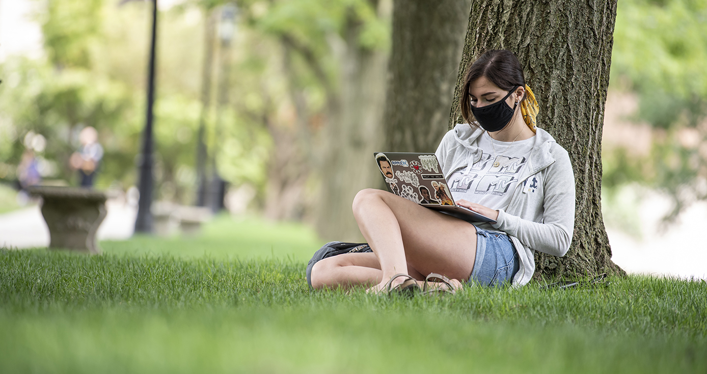 A person in a face mask, a sweater and shorts sitting by a tree, on a laptop