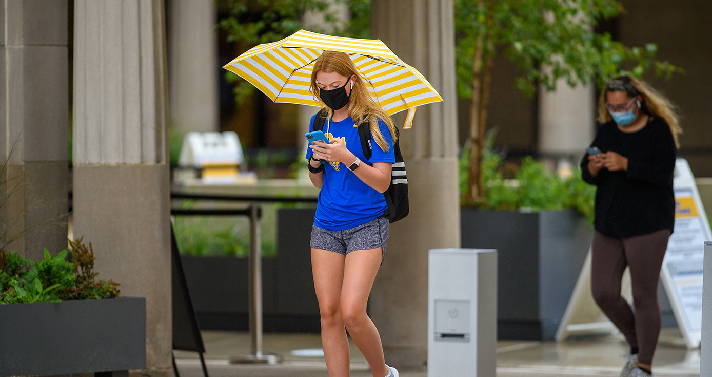 A student in a blue shirt walks down the sidewalk in a face mask and holding an umbrella and phone