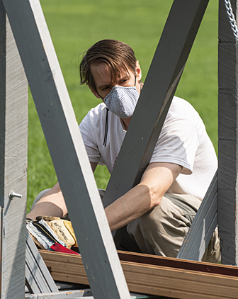 A person in a face mask squats next to a trebuchet