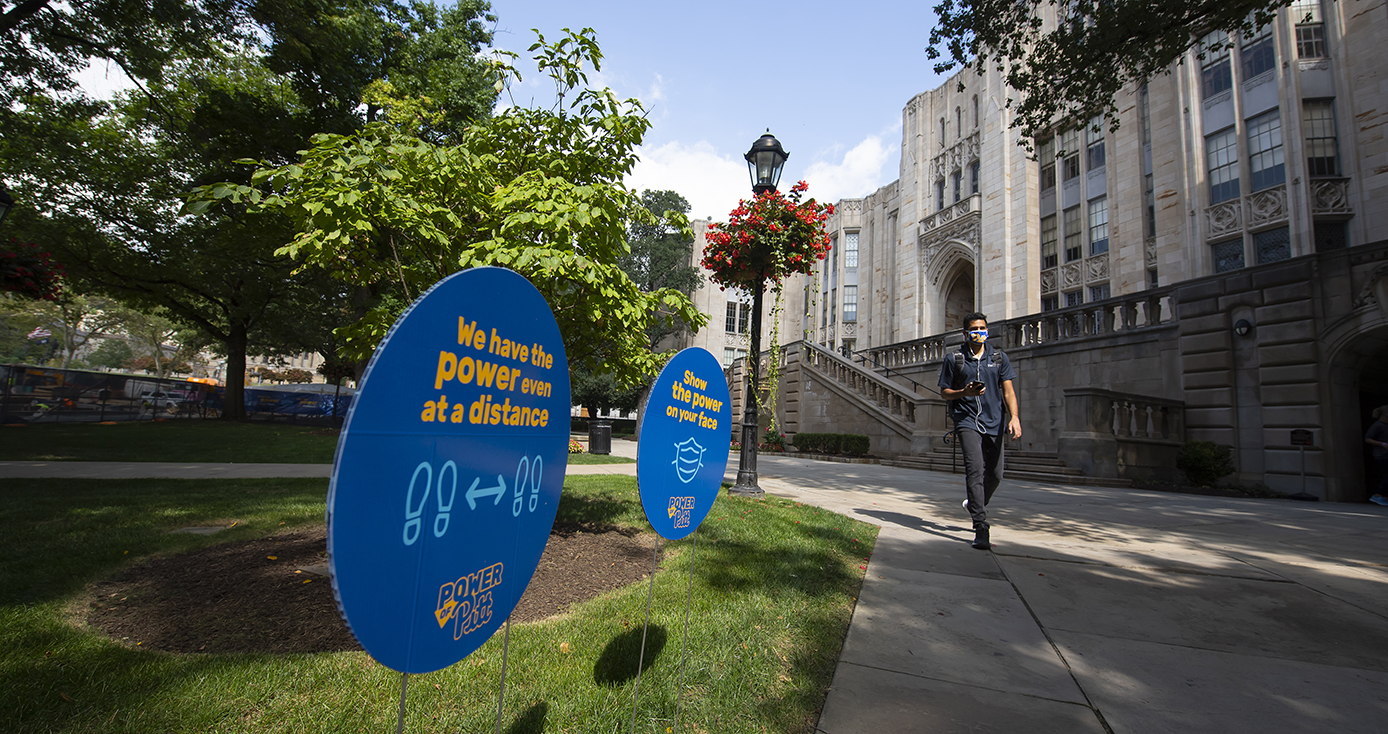 Two signs in the grass reminding students about the Power of Pitt, with a student in a face mask walking past