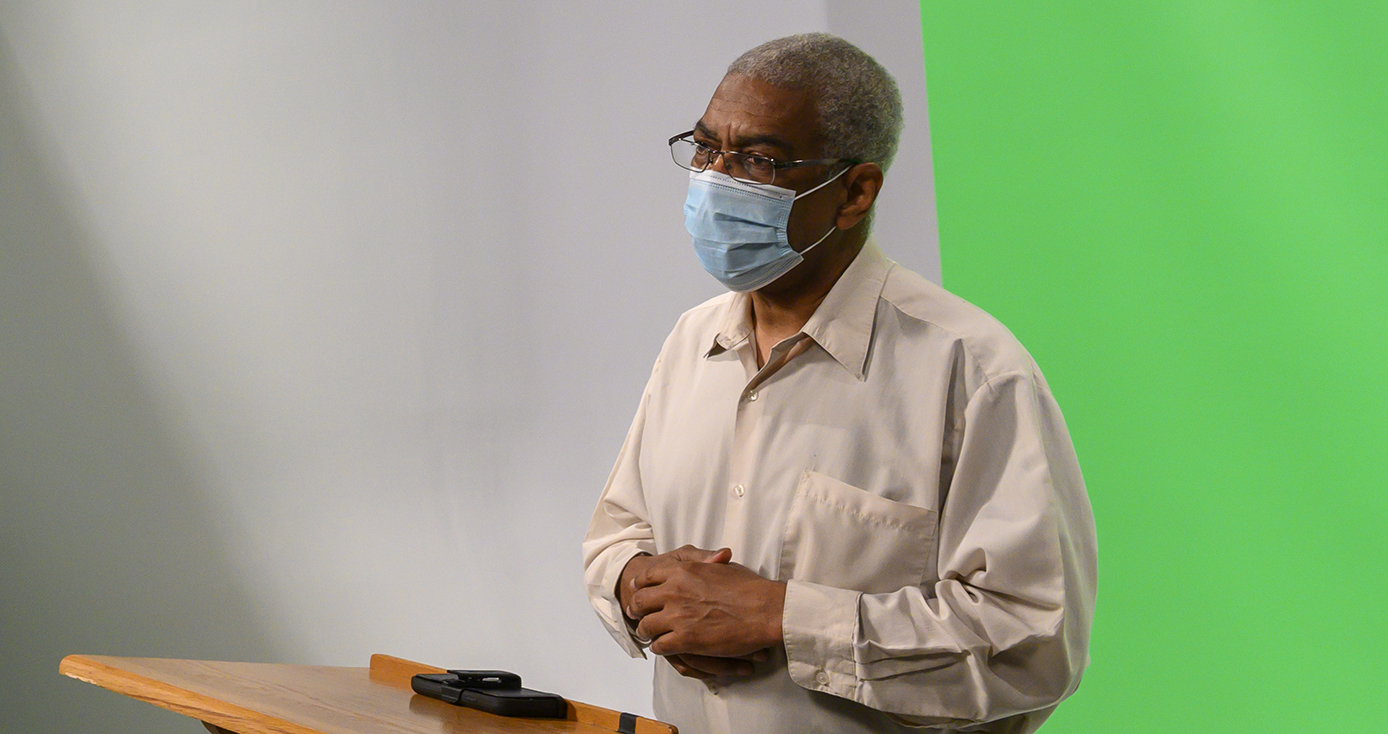 A person in a face mask and tan shirt stands in front of a green screen