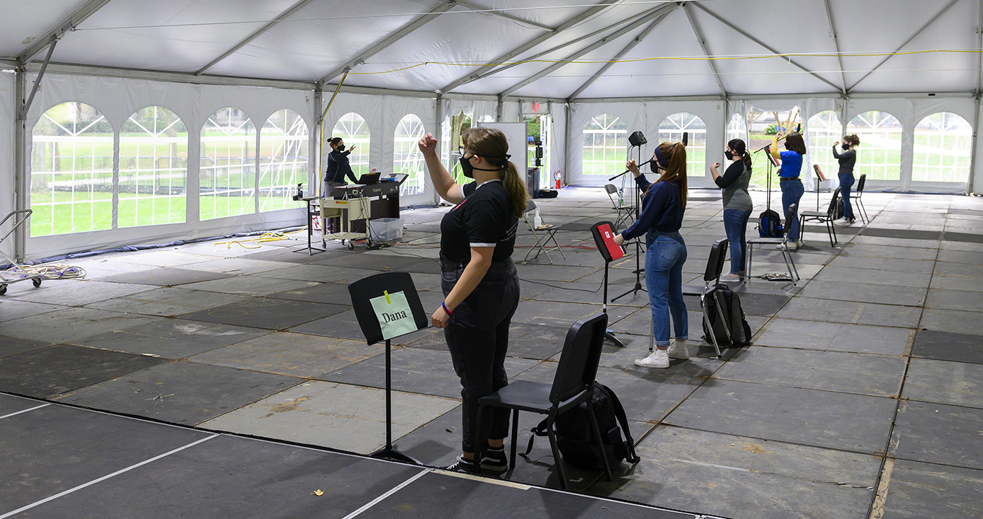 A woman in a face mask conducting to several students in face masks, inside a white tent