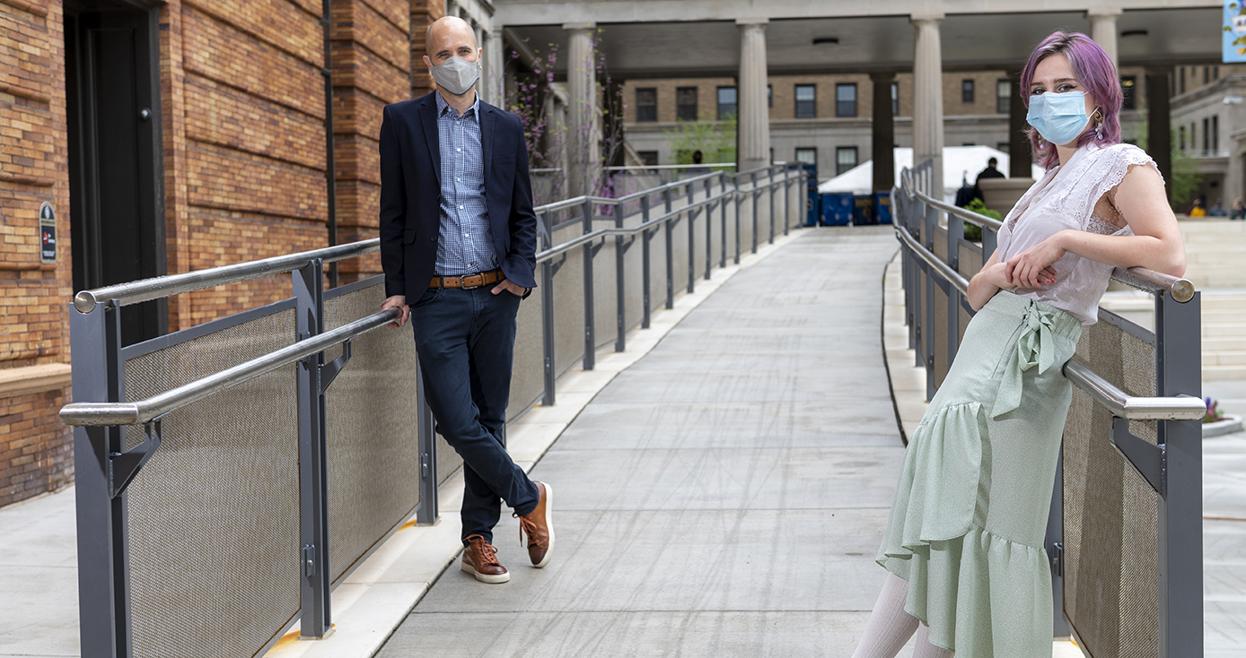 Dan Kubis leans against a rail on a walkway outside of a residence hall.  To the right is a woman wearing a mask.