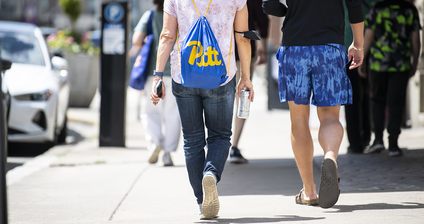 Two students walking down a sidewalk form behind, one wearing a blue Pitt bag