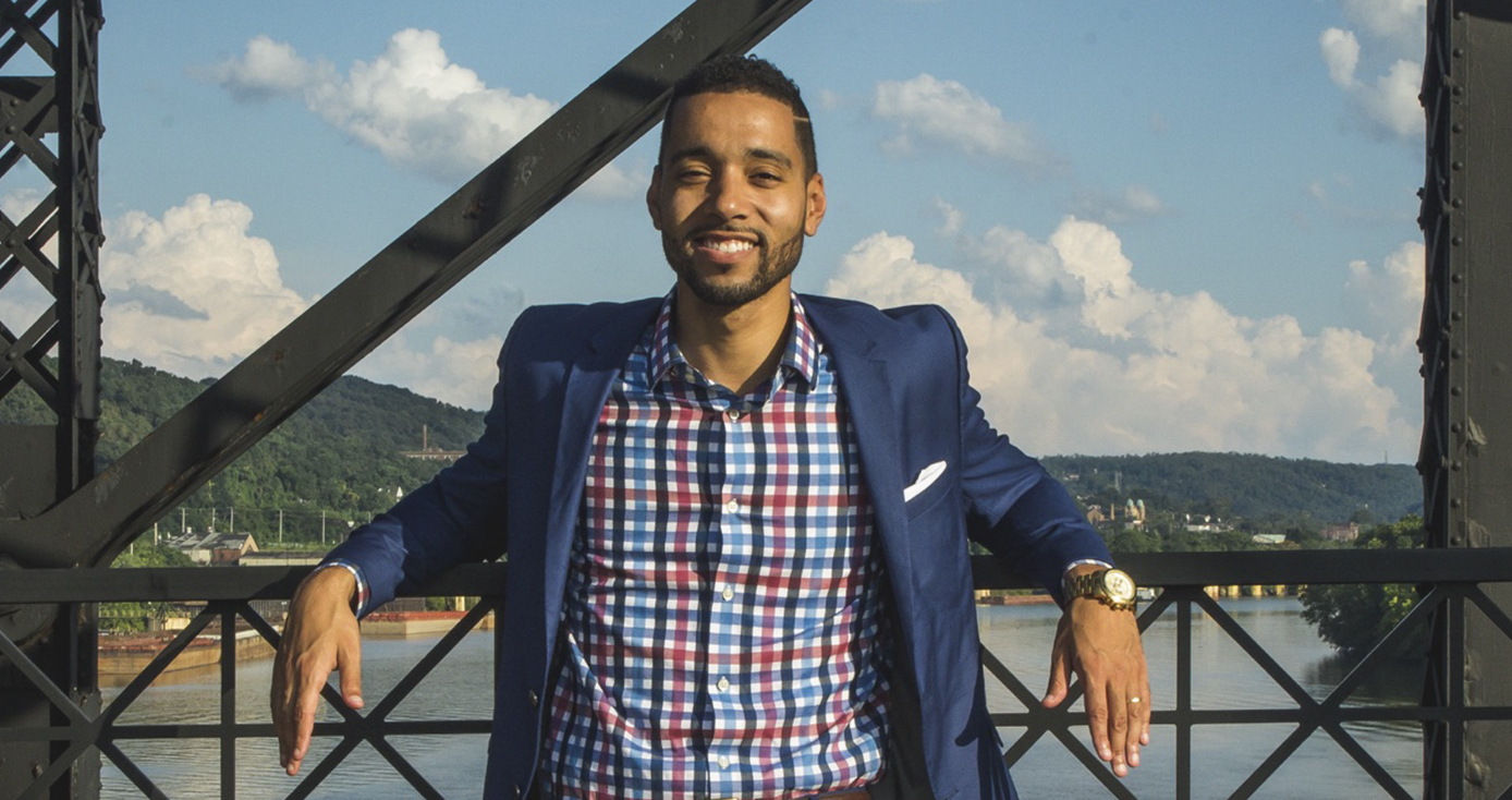 Brian Burley standing on a bridge over a river, elbows on railing, in checked shirt and blue jacket