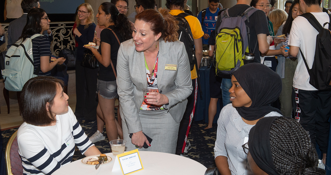 Belkys Torres speaking to students at a reception. Torres is wearing a gray top and skirt. 