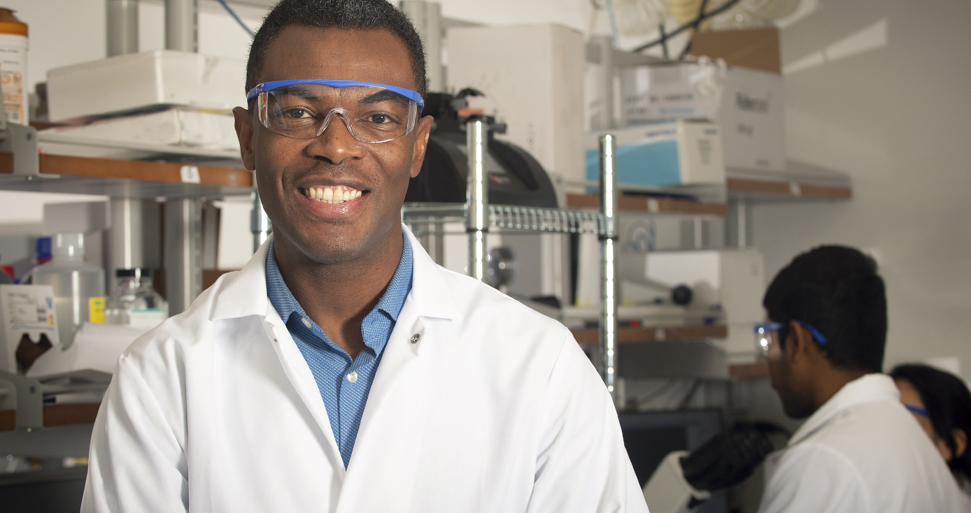 Portrait of Tagbo Niepa, in safety glasses, in a room with lab supplies on shelves in the background