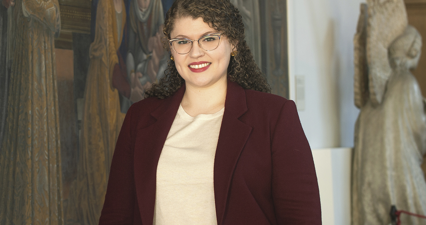 Conell in a burgundy blazer and white blouse, standing in front of a large piece of artwork