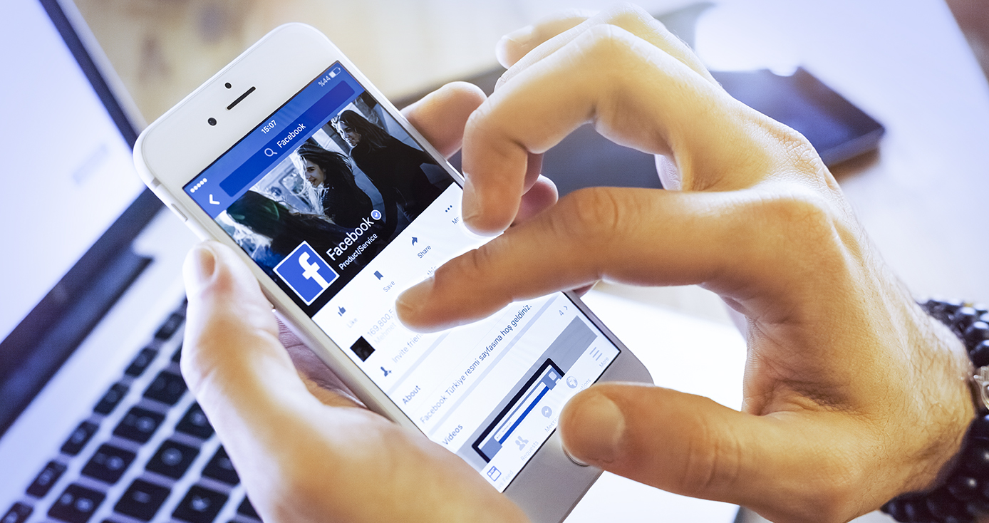 Close up of a person's hand holding a smart phone displaying Facebook, with a laptop computer in the background. 