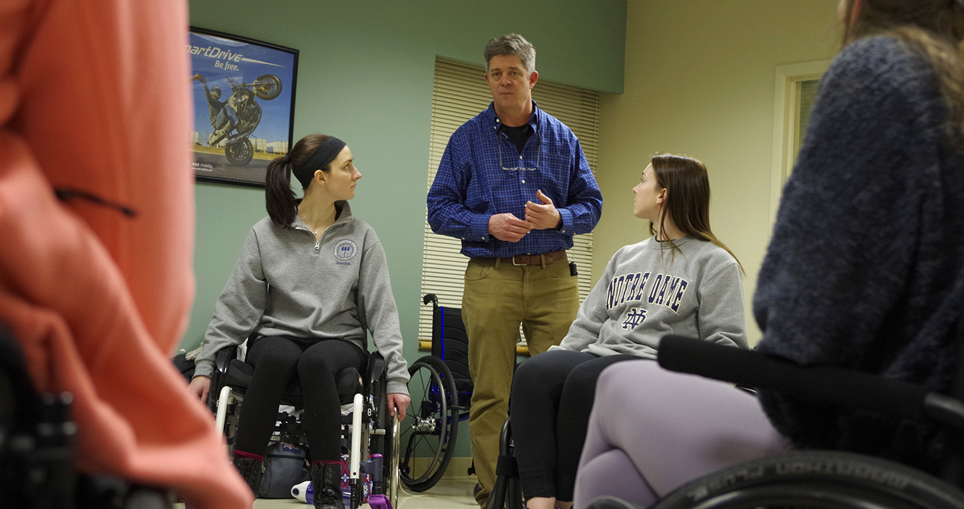 A person in a blue dress shirt and khaki pants speaking to four visible people in wheelchairs