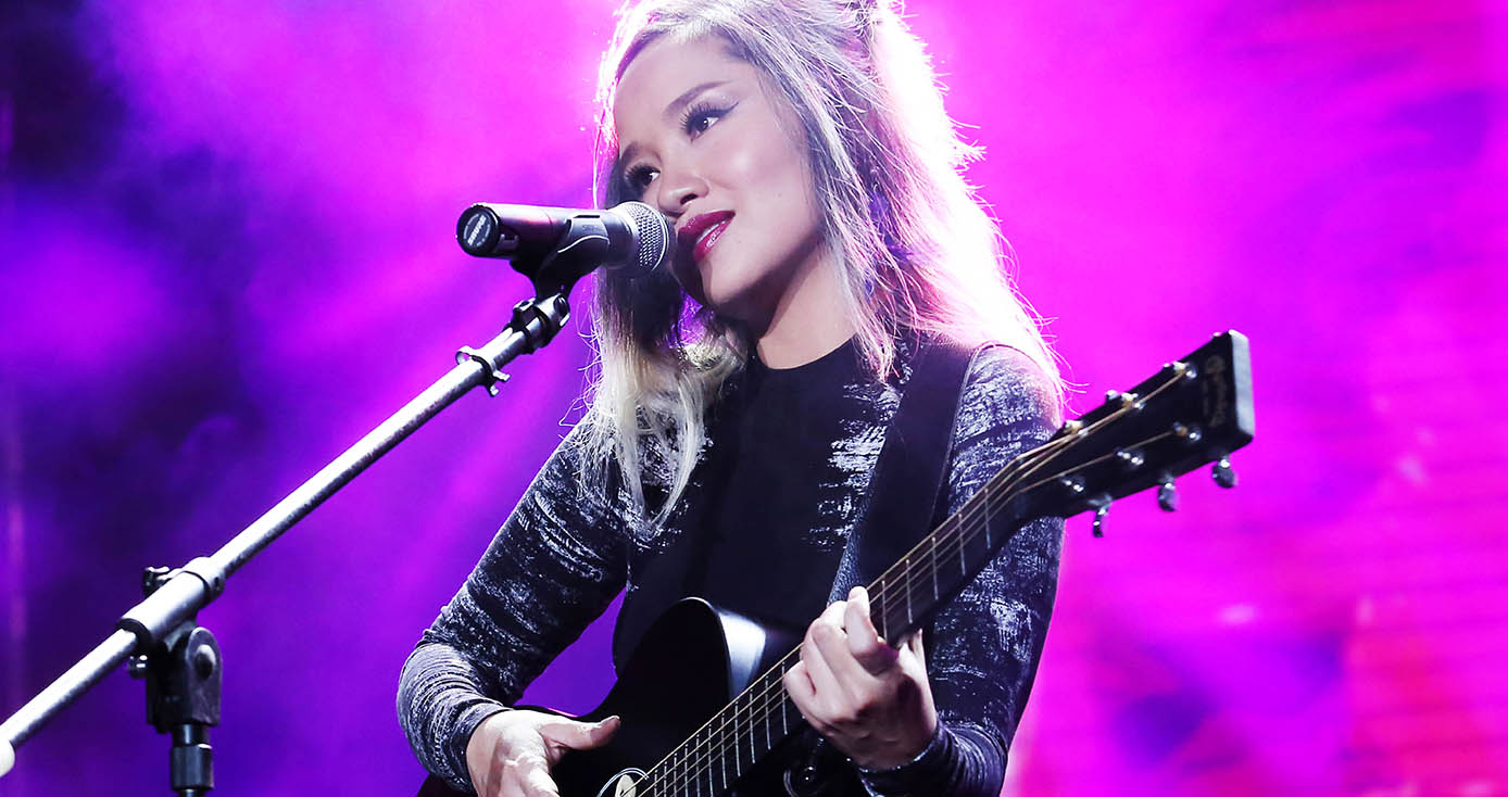 Khoi holding a black guitar in front of a neon pink background