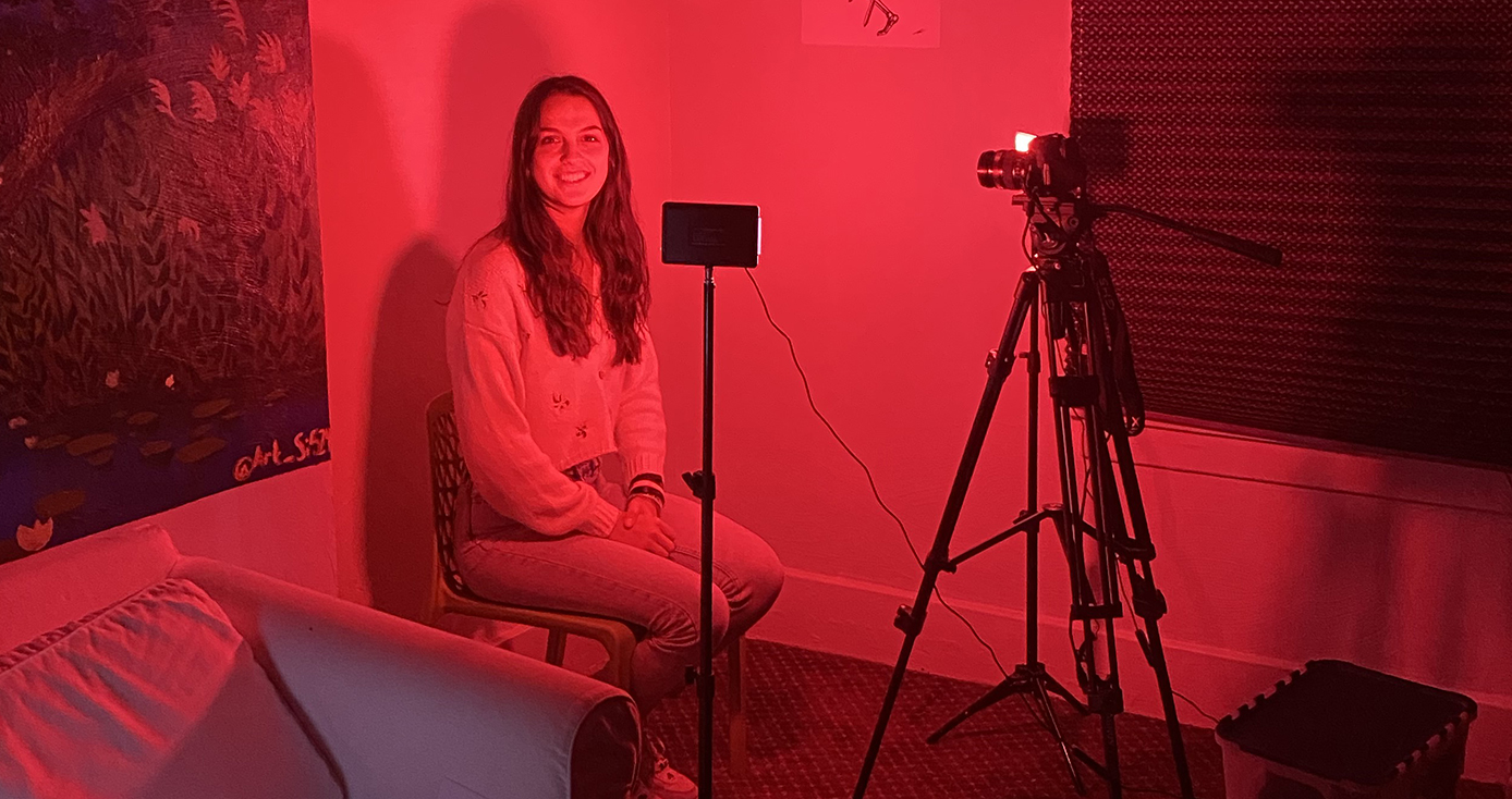 A person sitting in the corner of a dark red room with a camera facing them