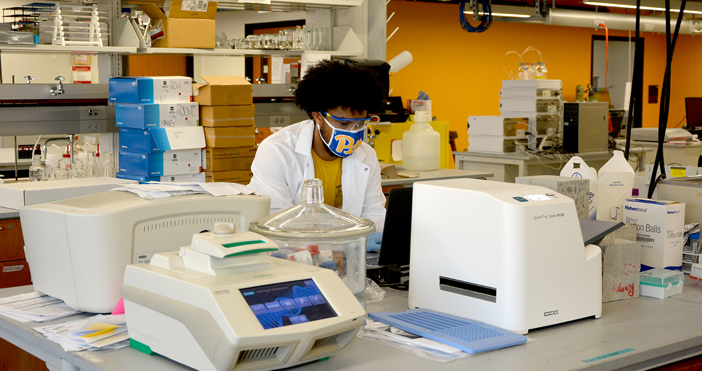 A person in a Pitt face mask and white lab coat works in a lab