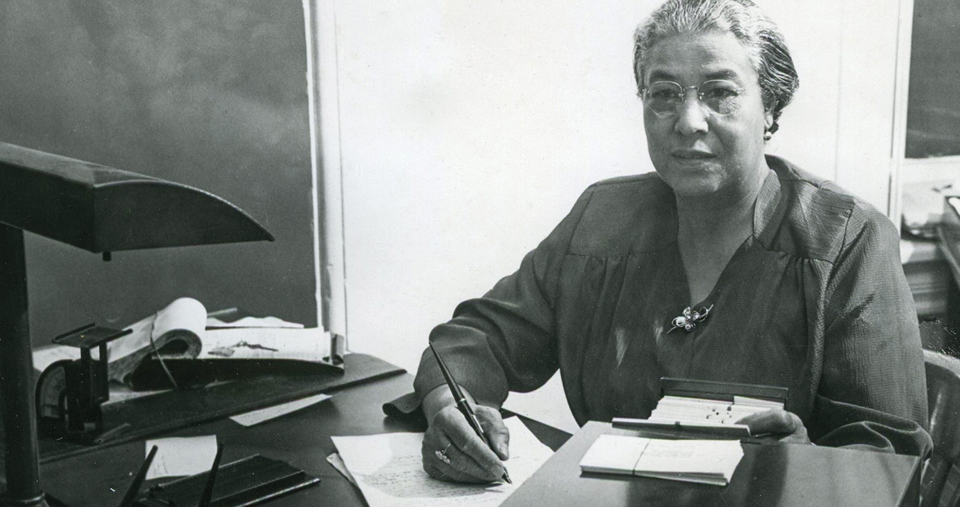 Ella P. Stewart at a desk, writing on a paper, in a black and white photograph. 