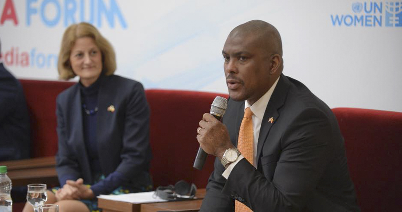 Hogan at a table on stage, holding a microphone. He's wearing a suit with an orange tie.