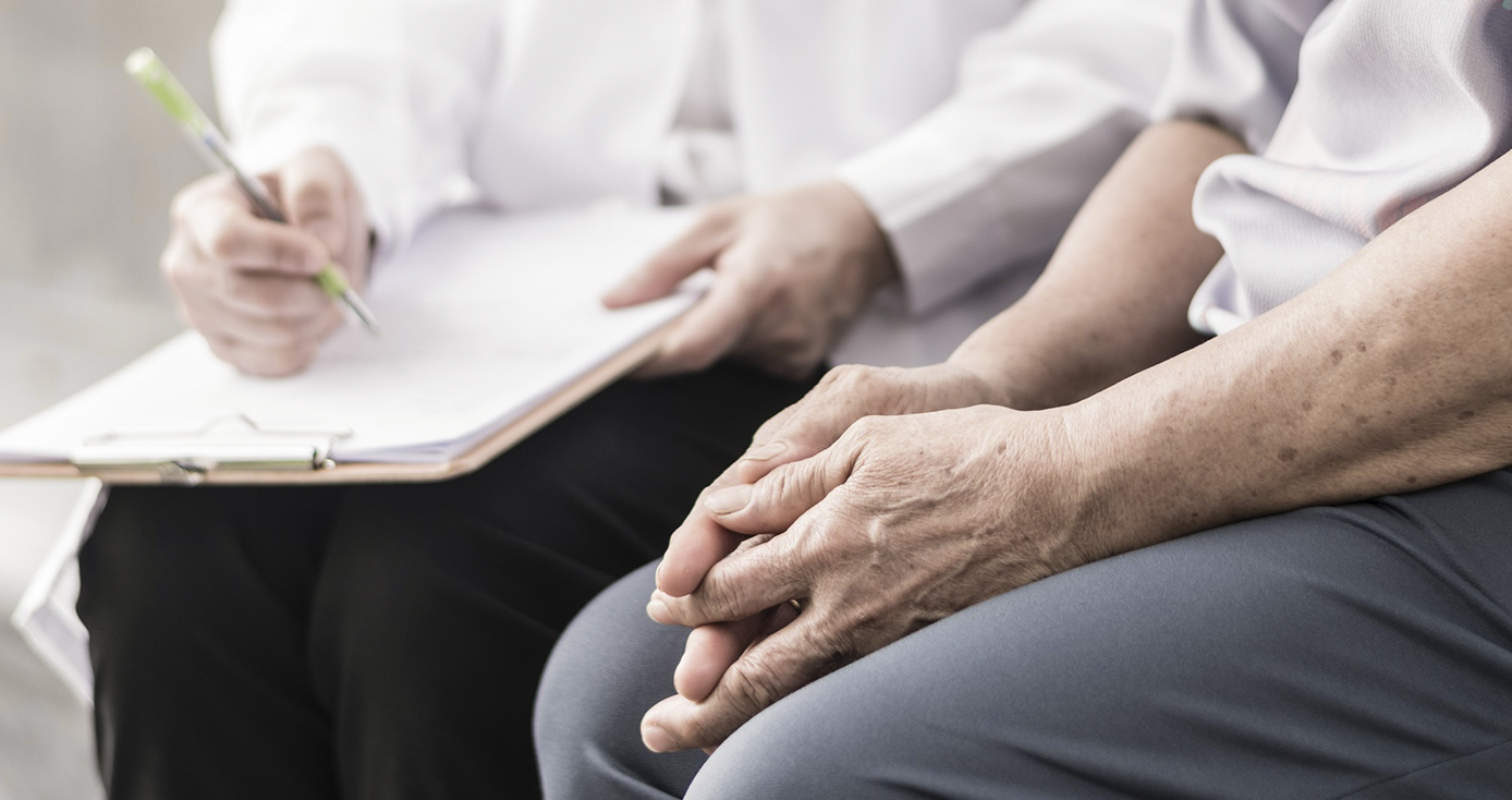 A person sits with hands on their lap, next to another person writing on a clipboard.