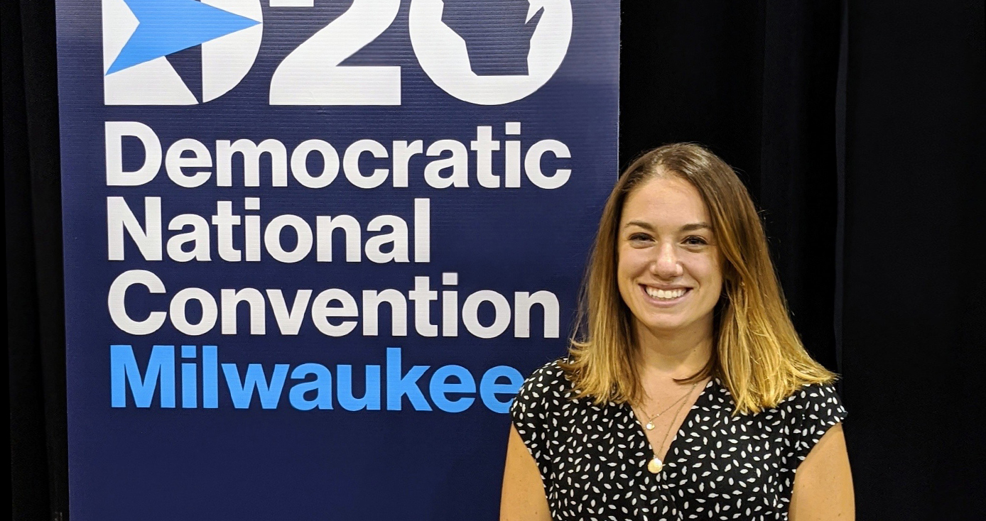 A woman in a black and white dress stands next to a DNC Milwaukee banner