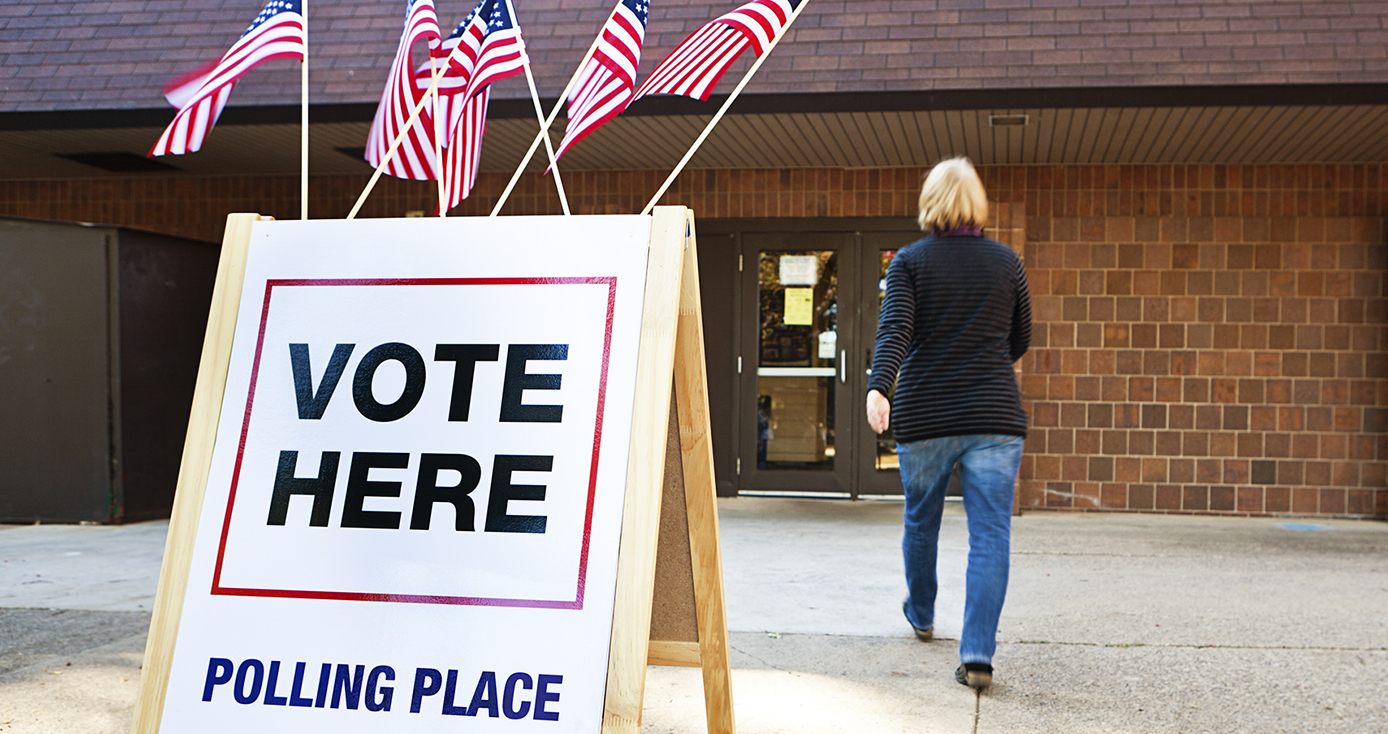 woman walking into a brick polling place building with a VOTE HERE sandwich board outside