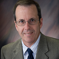 A man in a gray-brown jacket and a light collared shirt and dark tie