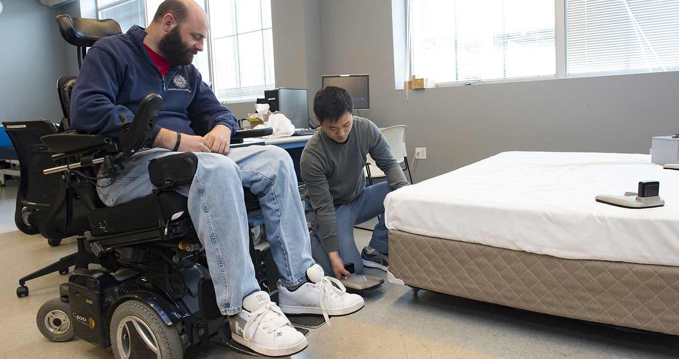 A man in a blue jacket and jeans in a wheelchair and a person in a gray shirt and jeans kneeling next to him