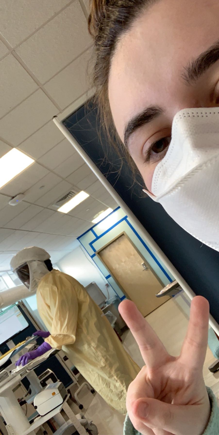 a woman in a mask making a peace sign with a medical professional in a full-body suit behind her