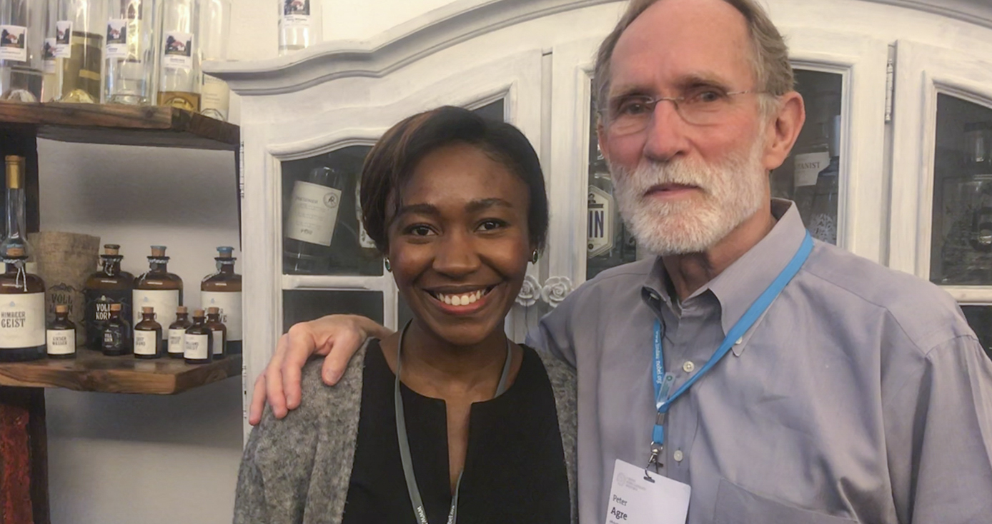 Olonisakin in a gray and black shirt, smiling, next to Peter Agre, a man with a gray beard, glasses, and wearing a bluish purple collar shirt