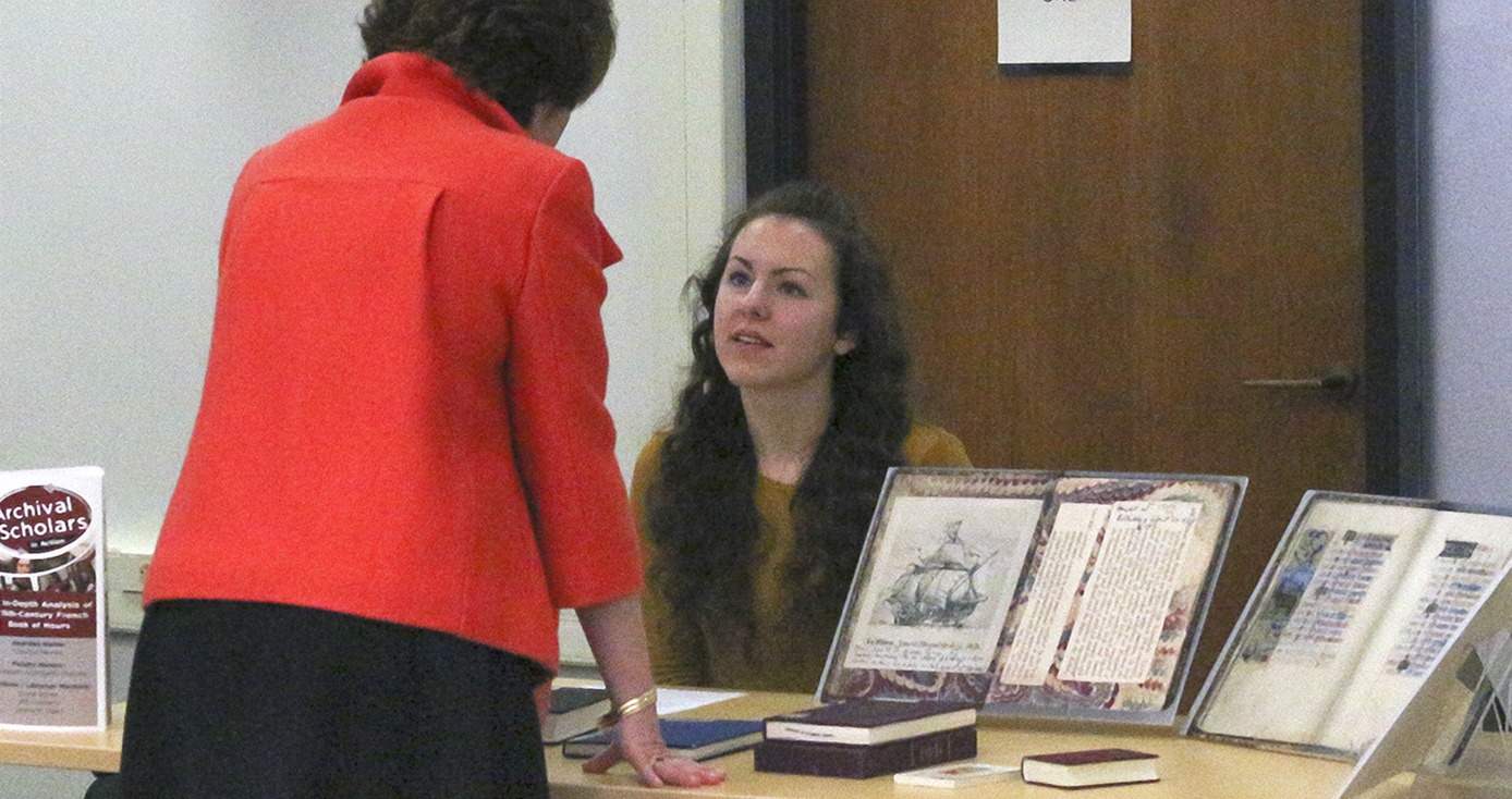 Claudia Haines seated as a table with her research project materials speaking with ULS library director