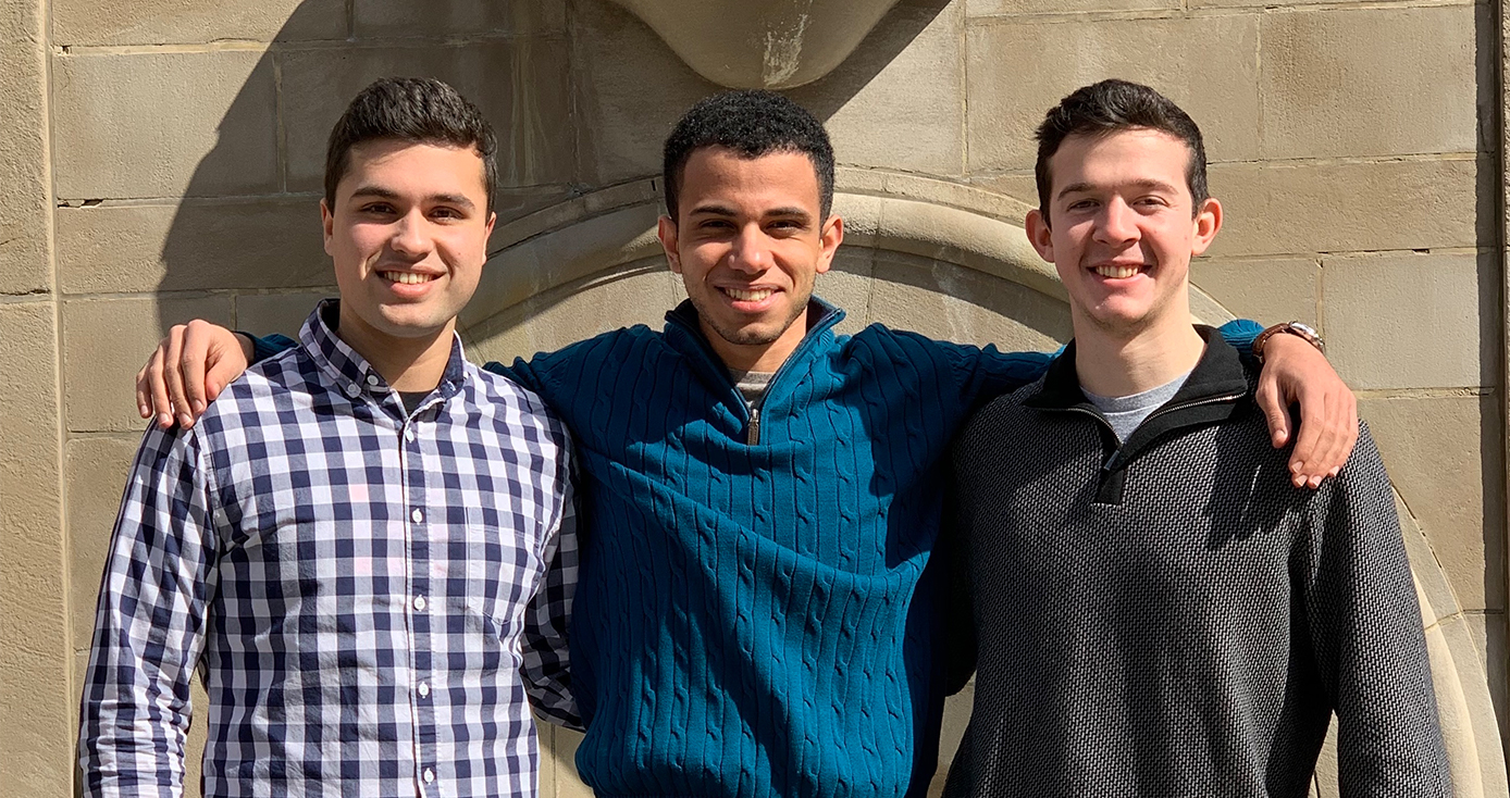 Three people standing together in dress shirts and sweaters against a building