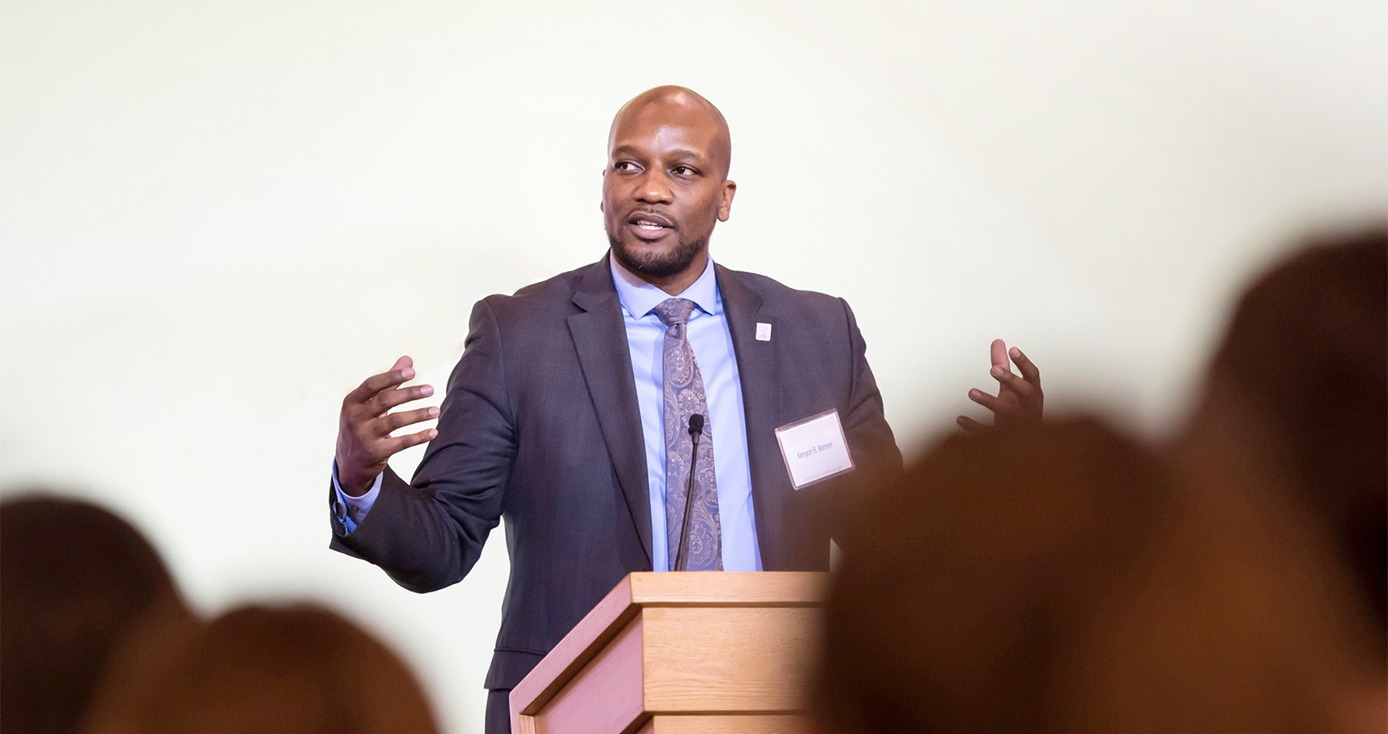 Kenyon Bonner speaking on a stage in a blue-gray suit and a collared shirt and tie