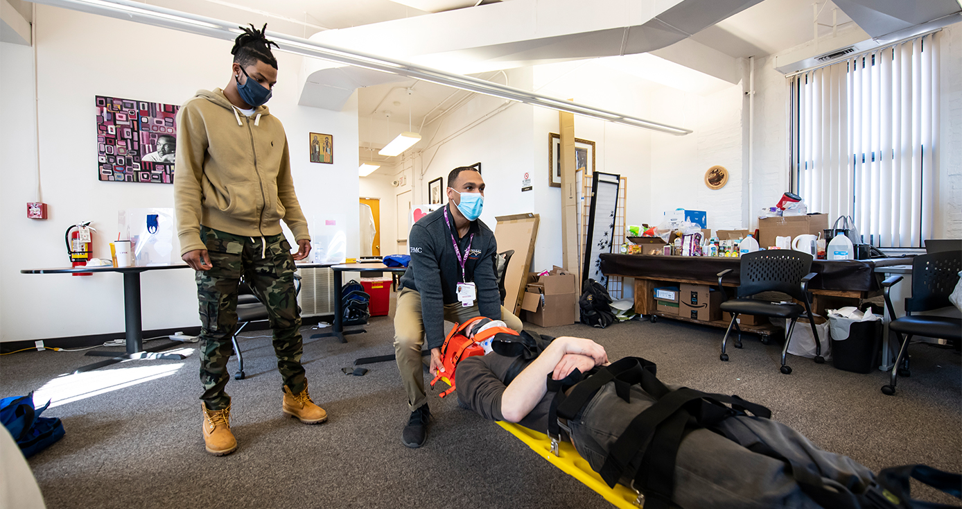 A person in a brown jacket and camouflage pants watches another person in a blue jacket lift a stretcher