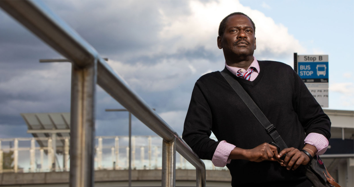 Brian Broome in a black sweater and pink shirt and a messenger bag leaning against a railing