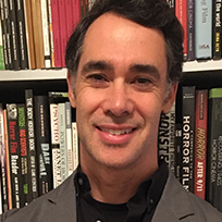 a man in a black shirt and gray jacket in front of a bookcase