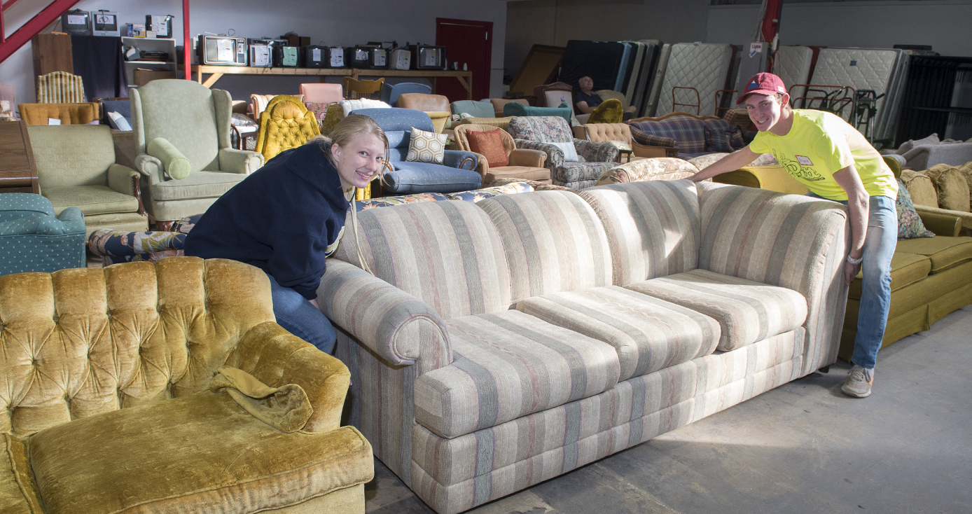 two students, a woman and a man, wearing neon yellow shirts and moving a couch