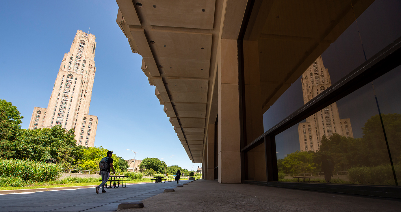 A student walks on campus, with the Cathedral of Learning in the background