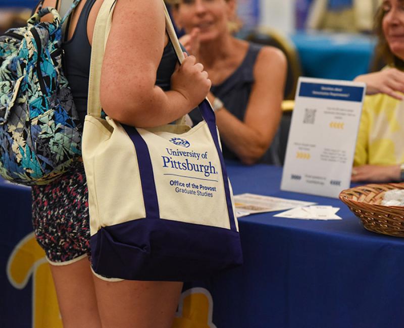 Person carries University of Pittsburgh tote bag while talking to women at a booth