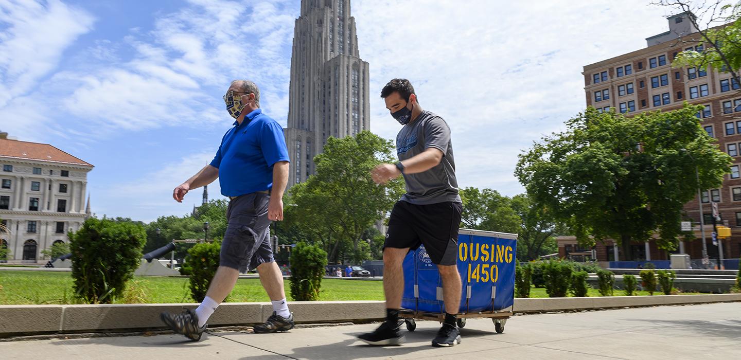Two men in masks walking with a housing cart with the Cathedral of Learning in the background