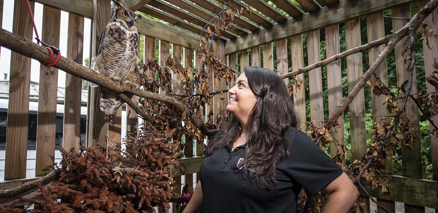 Katie Kefalos stands in an enclosure with an owl on a branch at her side