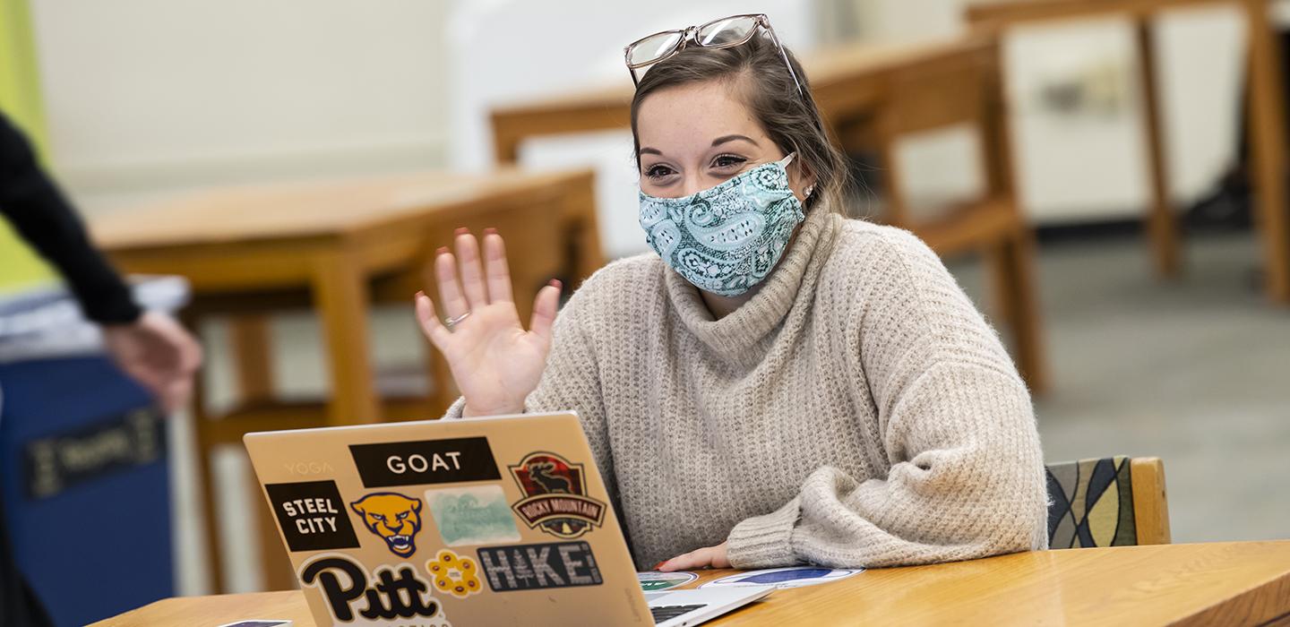 Student with mask using laptop, waving during video call