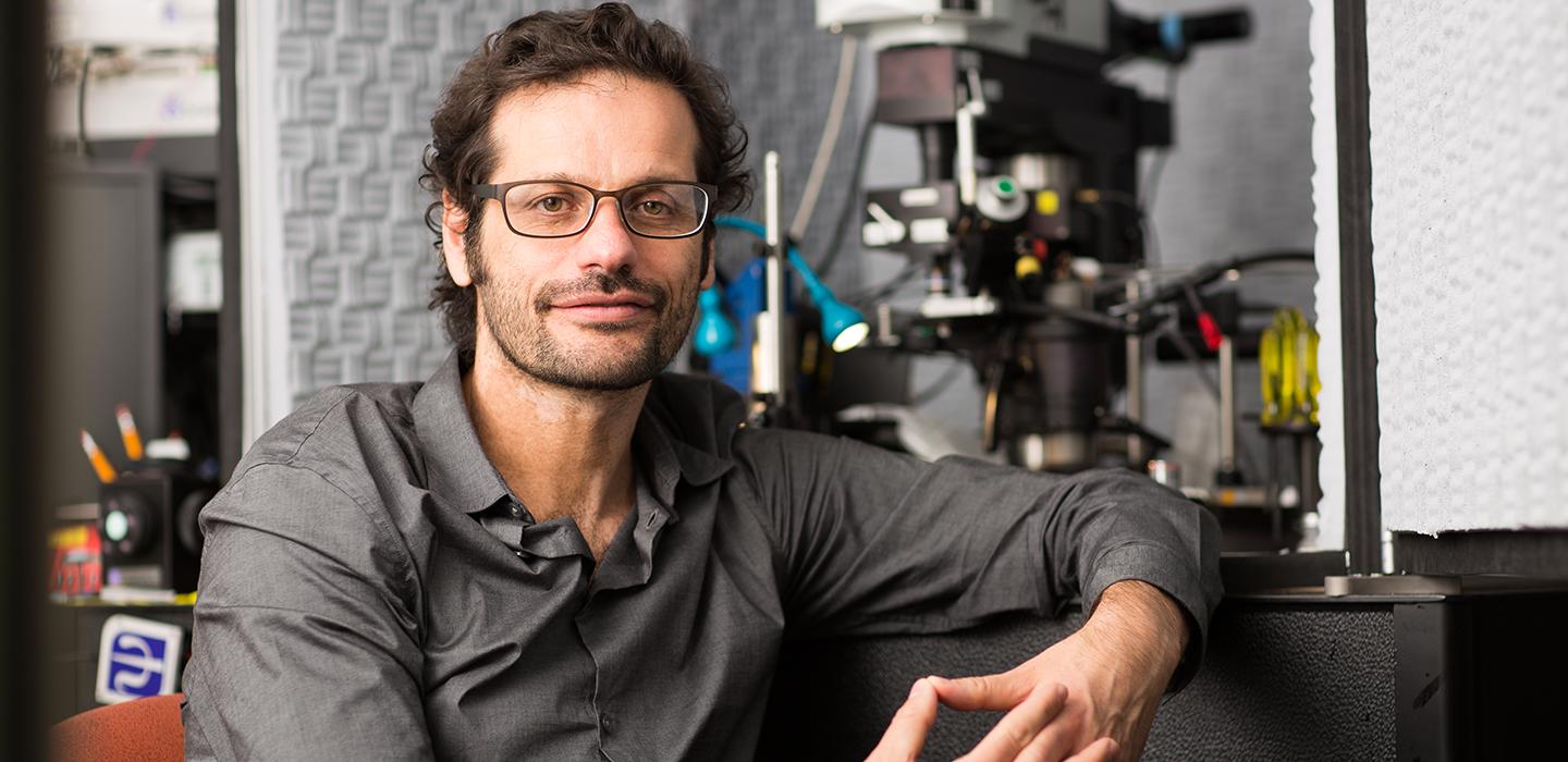 Thanos Tzounopoulos wearing grey shirt and glasses sitting on a chair