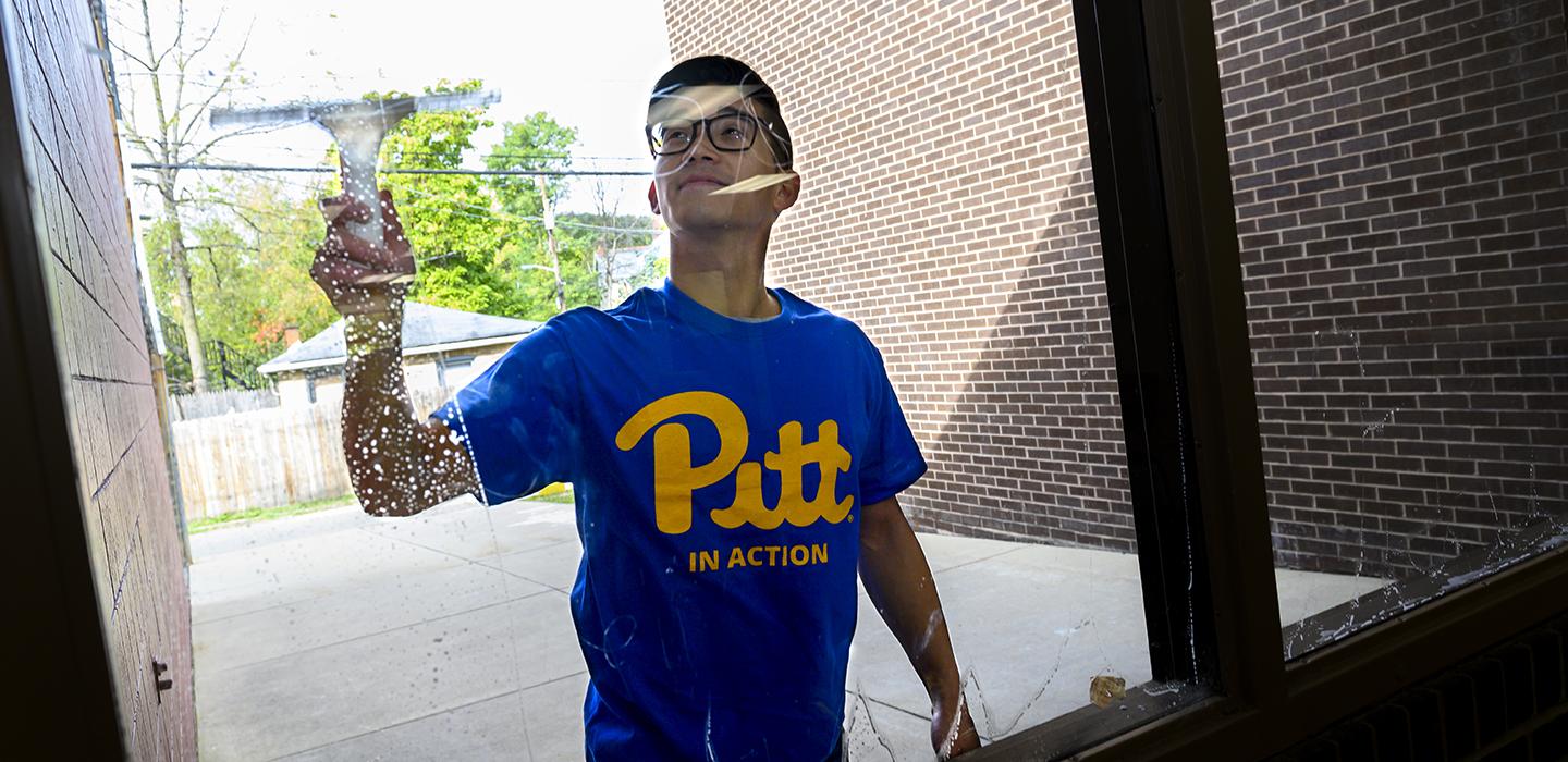 Man with blue Pitt shirt cleaning window from the outside