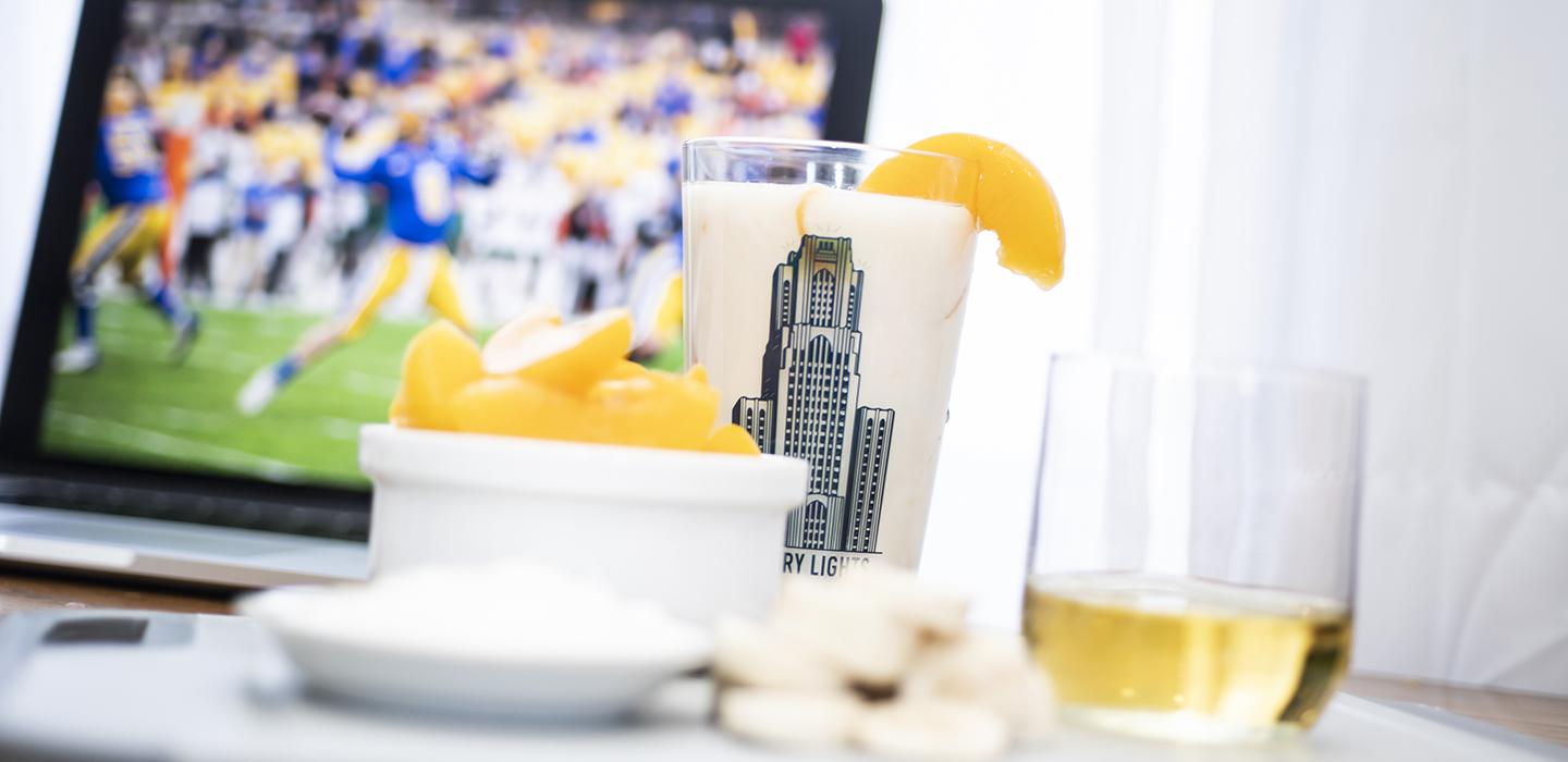 Peach smoothie with bowl of sliced peaches in front of laptop showing photo of Pitt football game