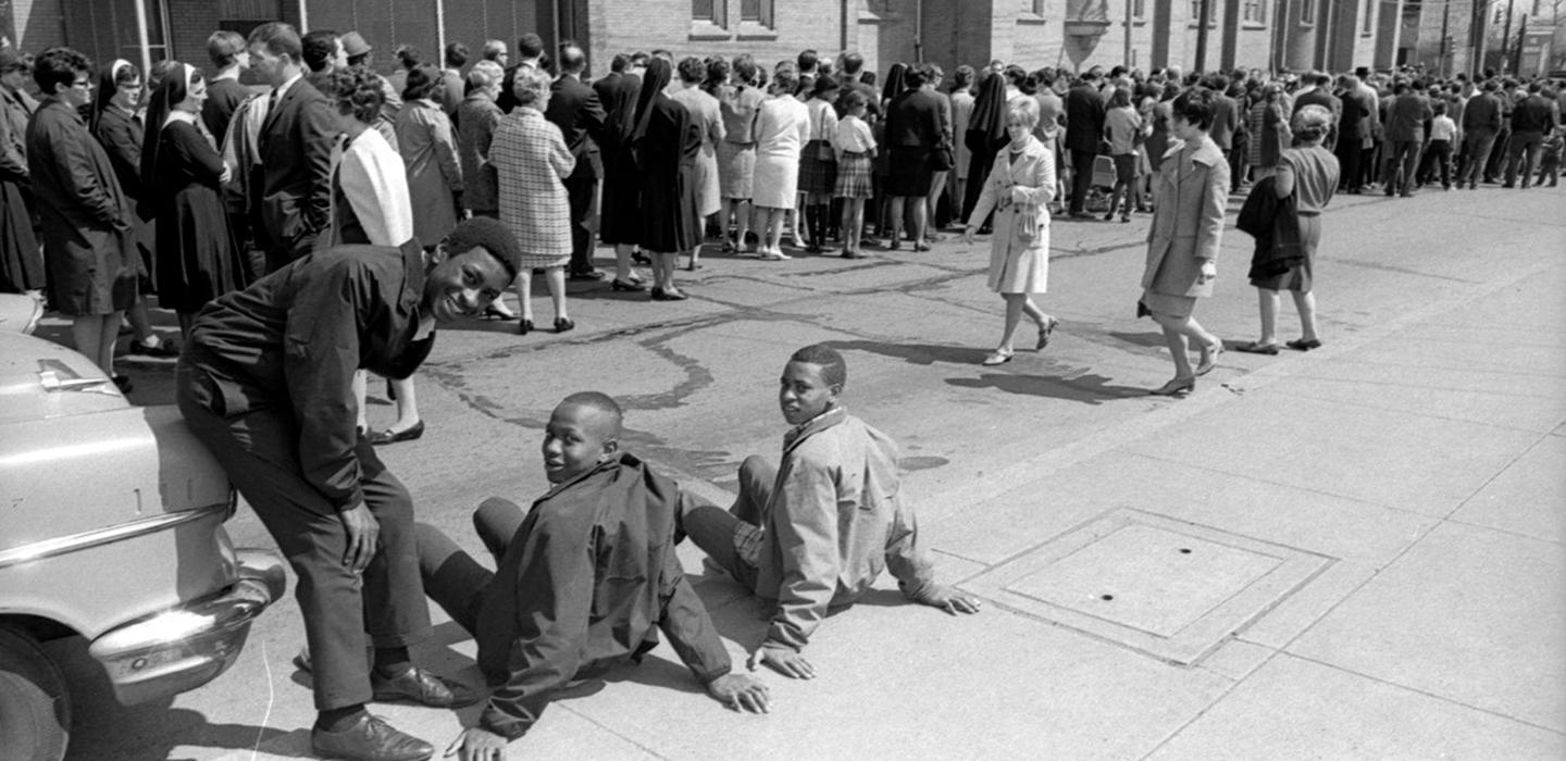 crowd of people gathered on street/sidewalk near a church with three boys in the foreground on National Day of Mourning following Martin Luther King Jr.'s assassination in 1968.